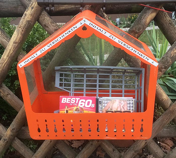 Orange Metallkiste an einem Zaun: „Inhalt zu verschenken“ mit CDs