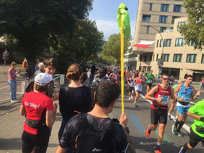 Zuschauer mit neonfarbener Mütze am hochgestreckten Zollstock sieht wartend dem Läufer:innenfeld entgegen