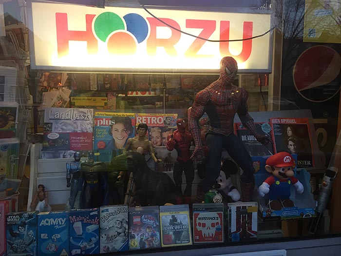 Schaufenster-Auslage eines Kiosks mit Magazinen, Playstation-Spielen und Figuren (Batman, Dinosaurier, Micky Maus, Super Mario und ein großer Spiderman)