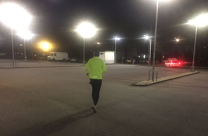 Läufer in neongelber Jacke auf einem großen Parkplatz im Morgendunkel mit grellweißen Laternen