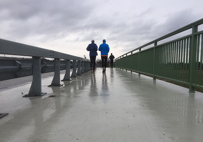 Drei Läufer auf einer Brücke, links die Leitplanke, rechts ein grünes Geländer, darüber ein wolkenverhangener Himmel