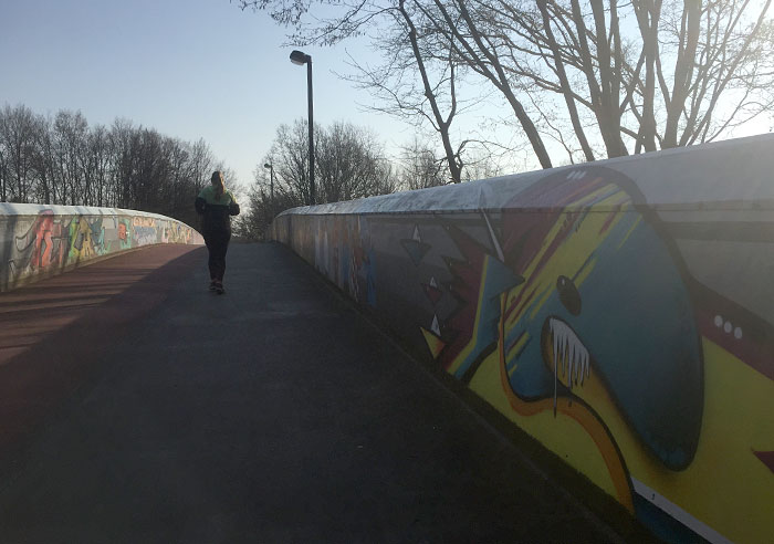 Läuferin auf einer Autobahnbrücke, deren Metall-Seitenbrüstungen mit bunten Graffitis besprüht sind