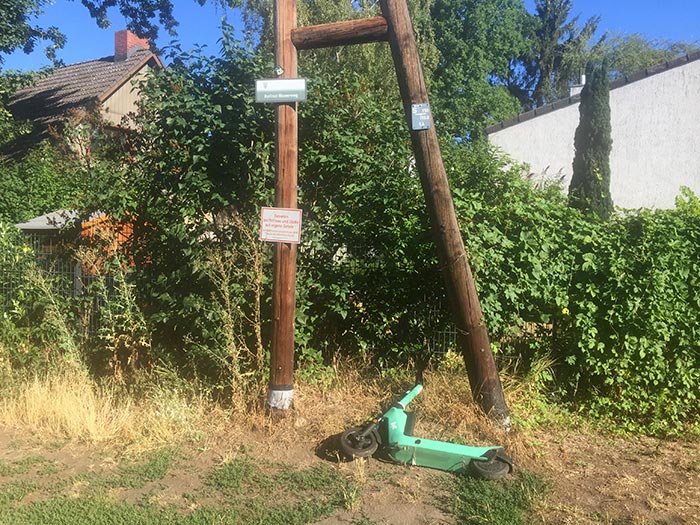 Alter hölzerner Stromleitungsmast mit Schild „Berliner Mauerweg“, davor liegt ein E-Scooter im Gras
