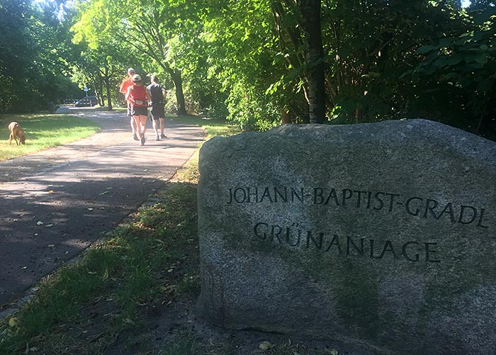 Im Vordergrund großer Findling mit aufschrift „Johann-Baptist-Gradl-Grünanlage“, im Hintergrund Laufgruppe auf Parkweg