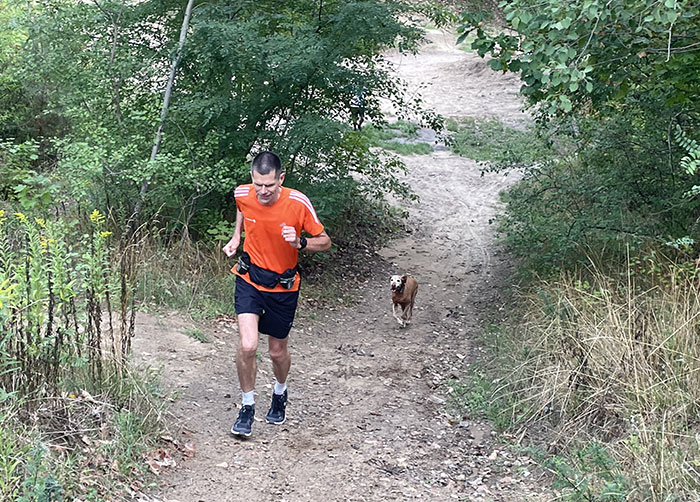 Läufer im orangen Shirt läuft einen sandigen Anstieg hoch, gefolgt von einem Hund