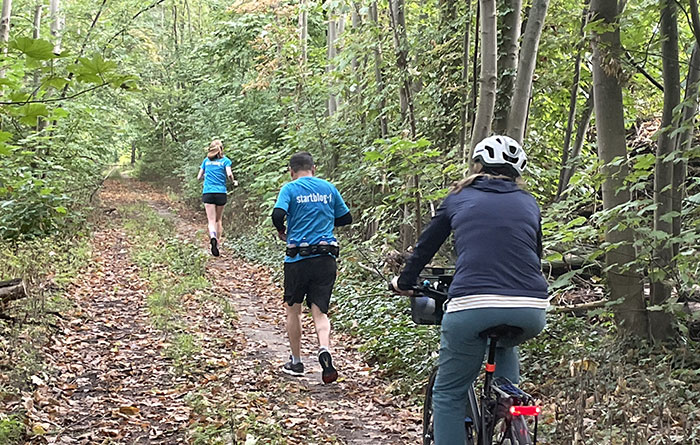 Läuferin und Läufer in blauen Laufshirts, begleitet von Radfahrerin auf einem schmalen Waldpfad