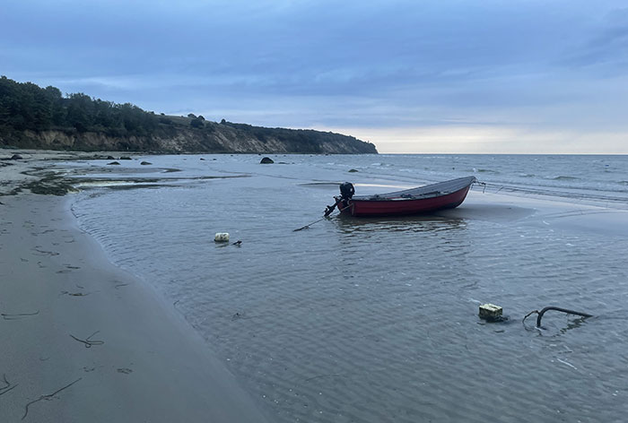 Kleines rotes Boot mit Außenbordmotor liegt im seichten Gewässer des Strands, im Hintergrund die Küste
