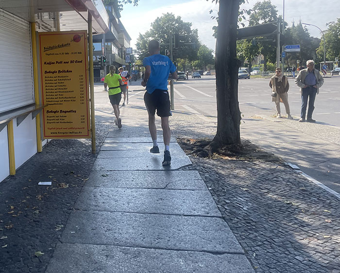 Läuferin in neongelbem Shirt und Läufer in hellblauem Shirt biegen an einem Imbiss um die Straßenecke