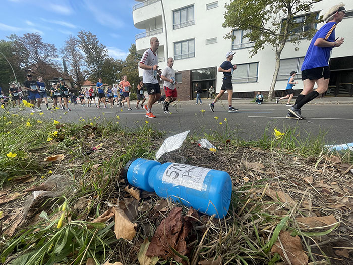 Im Vordergrund eine weggeworfene Trinkflasche eines Marathonläufers, im Hintergrund das Läuferfeld