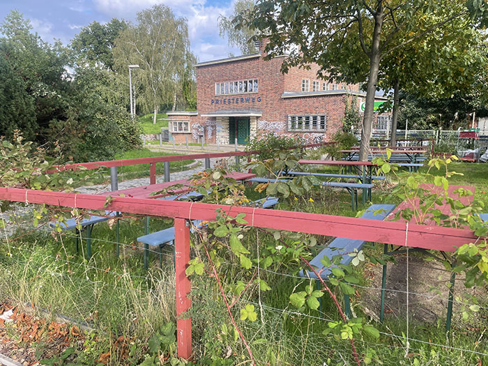 Blick über Sommergarten-Bänke und rankende Pflanzen auf das alte Gebäude des S-Bahnhof Priesterweg