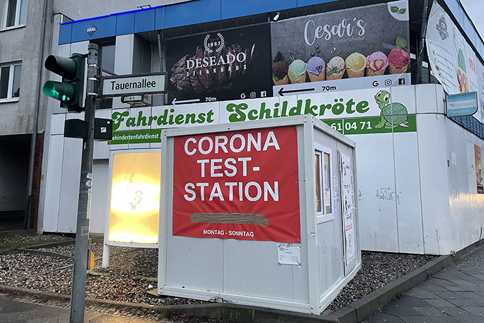 Mini-Container mit Aufschrift „Corona Teststation“ vor vielen anderen Werbeschildern an der Fassade dahinter