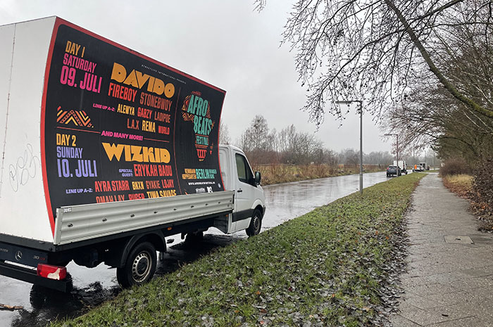 Einsame Straße im Nieselregen, am Straßenrand ein Transporter mit Werbeaufbau, auf dem in bunter Typografie Konzerte im nächsten Jahr beworben werden