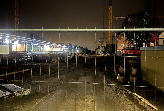 Blick durch einen Metall-Bauzaun auf eine Baustelle mit erleuchtetem neuen S-Bahnhof