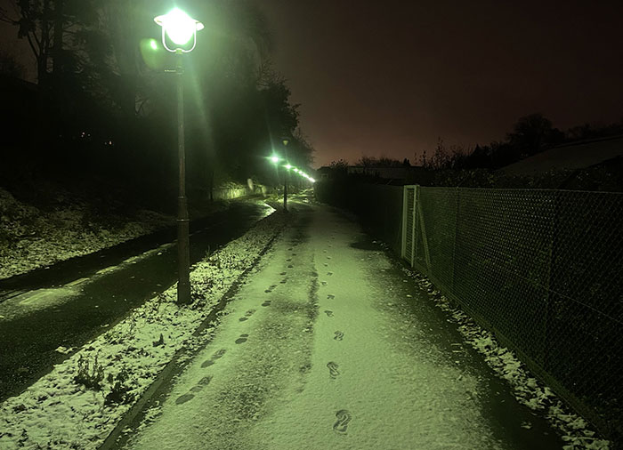 Von Laternen erleuchteter Fuß- und Radweg in der Morgendunkelheit, auf einem dünnen Schneebelag sind Fußspuren zu sehen
