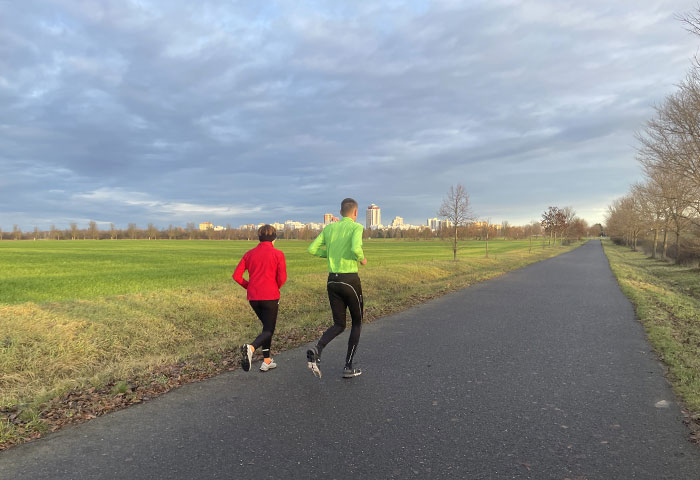 Läuferin und Läufer auf breitem Asphaltweg, am Horizont die Silhouette eines Hochhausgebiets