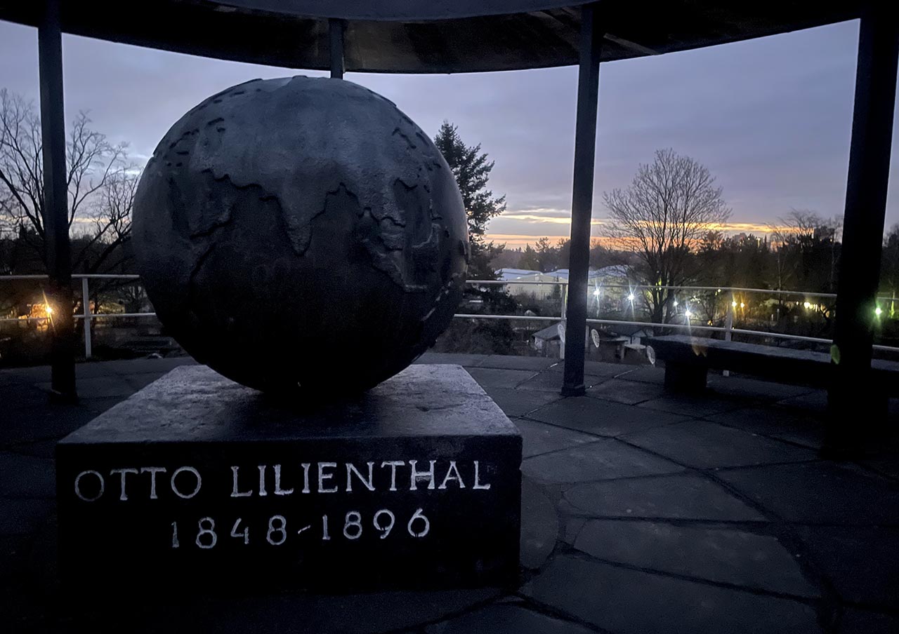 Auf dem Lilienthal-Denkmal: Große metallene Weltkugel auf einem Sockel, auf dem „Otto Lilienthal 1848-1896“ steht, im Hintergrund Lichter der umgebenden Häuser