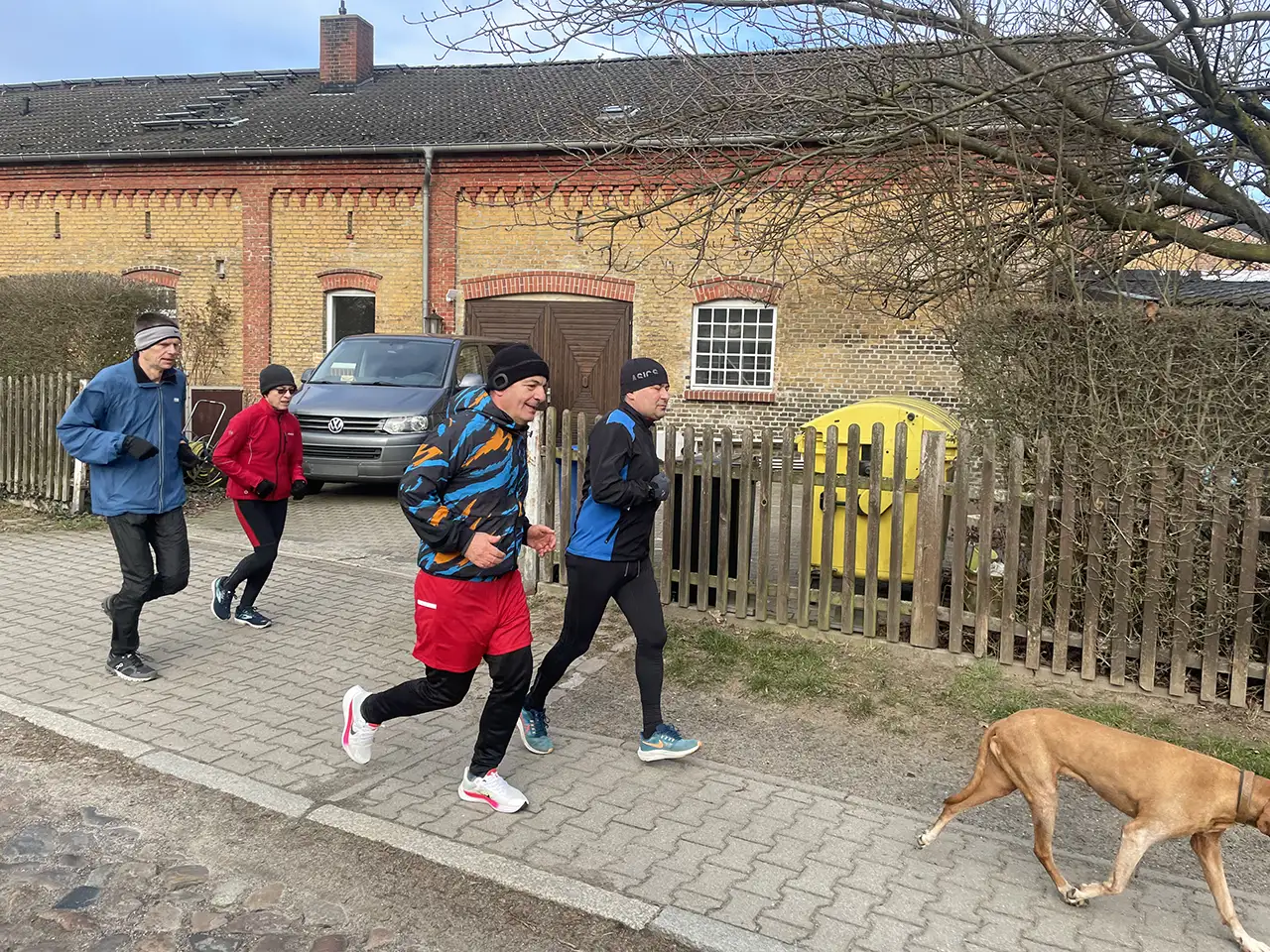 Laufgruppe mit Hund vor einem bäuerlichen Backsteingebäude mit gelben und roten Ziegeln