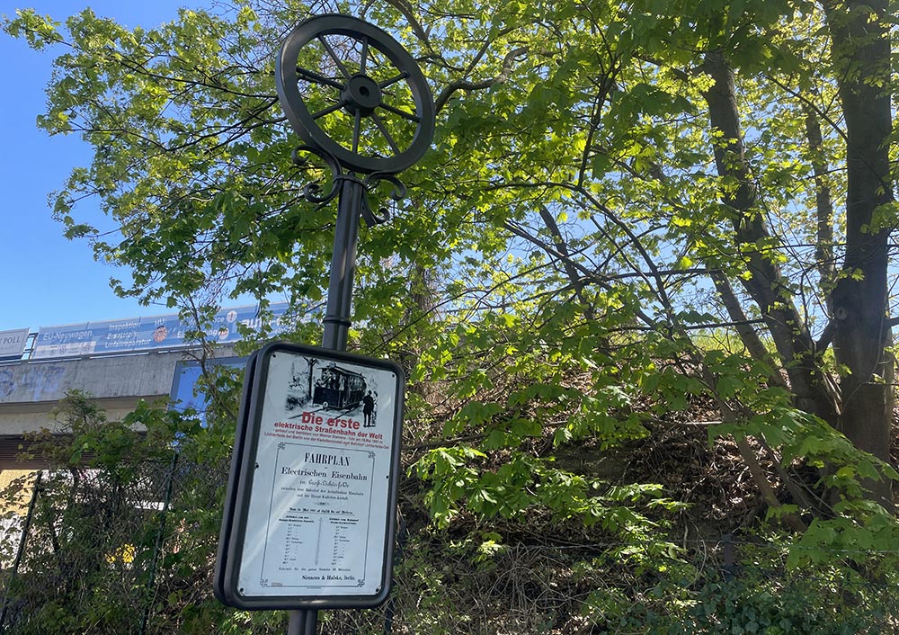 Alter Laternenpfahl, der oben in einem gusseisernen Rad endet und ein Schild trägt mit Hinweis auf die erste elektrische Straßenbahn der Welt