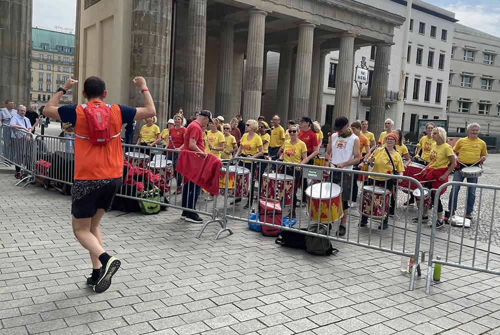 Trommelgruppe in gelben Shirts am Brandenburger Tor, ein vorbeilaufender S25-Teilnehmer reckt die Hände hoch