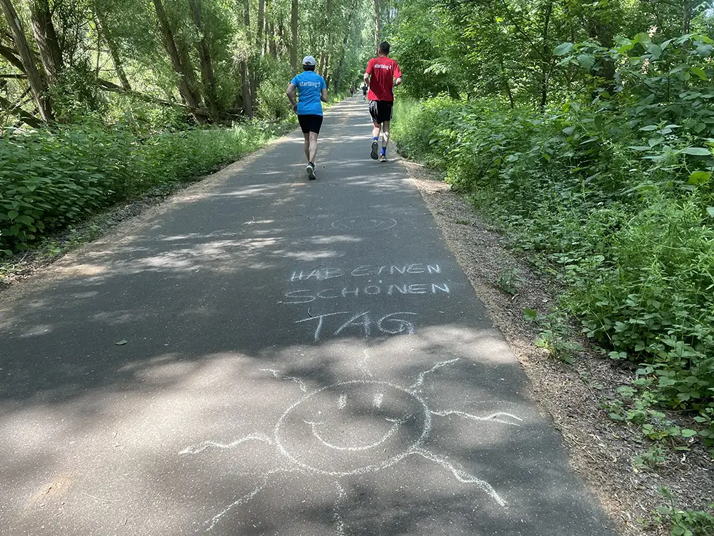 Läuferpaar auf Asphaltweg, auf den jemand mit Kreide eine Sonne gemalt hat und den Spruch „Hab einen schönen Tag“