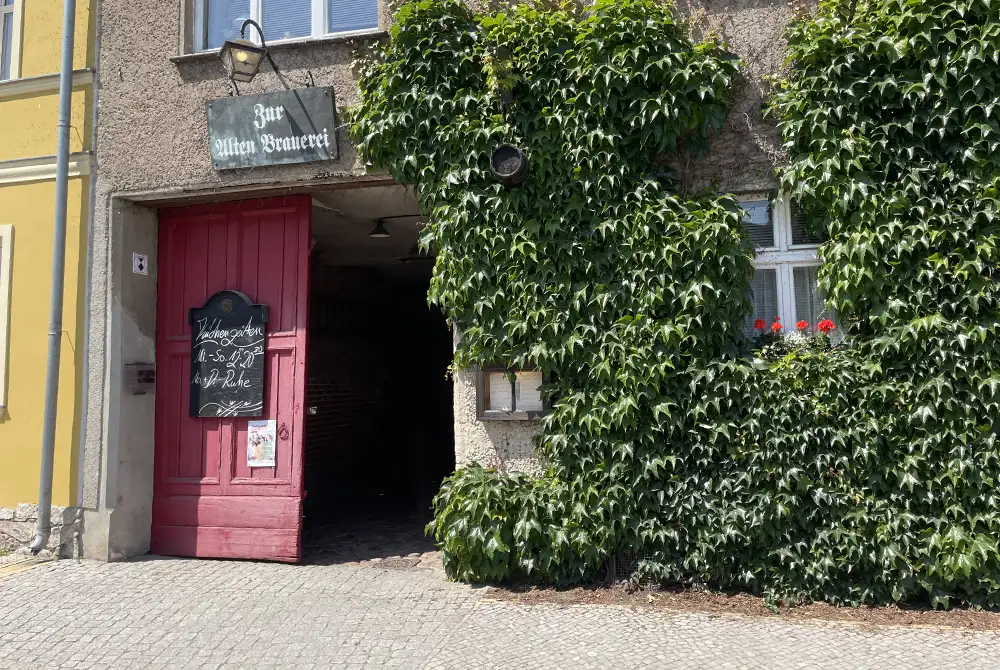 Efeuberankte Fassade mit rotem Holz-Eingangstor, darüber das Schild „Zur Alten Brauerei“