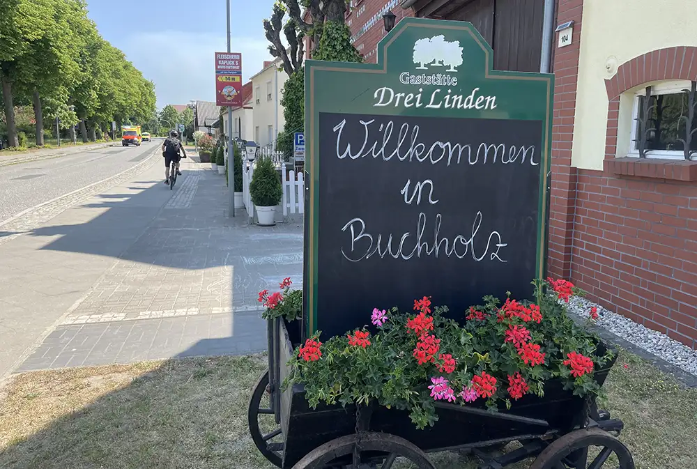 Mit Kreide beschriftete Tafel in einem Holzkarren mit Blumen an einer Gaststätte „Willkommen in Buchholz“