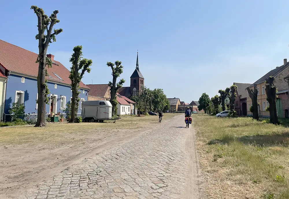 Schmale Kopfsteinpflaster-Straße durch breite Dorfschneise, im Hintergrund ein Kirchturm