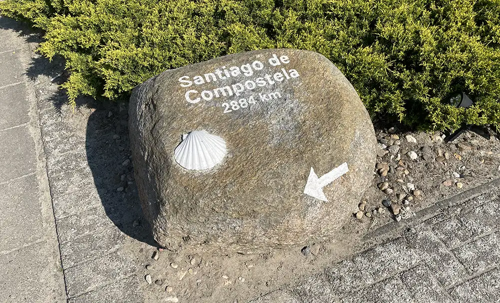 Feldstein im Vorgarten mit weißer Muschel und Pfeil sowie Beschriftung Santiago de Compostela 2884 km
