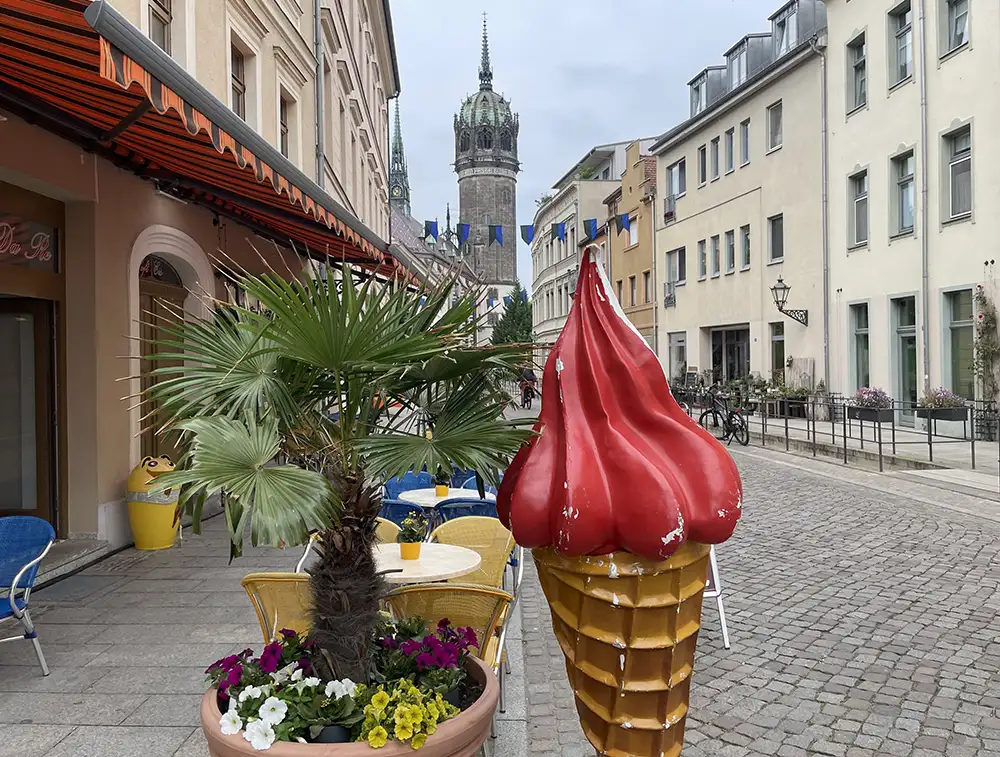 Große Plastik-Eiswaffel neben Palmen-Topf vor einem Eis-Café, im Hintergrund der Turm der Schlosskirche