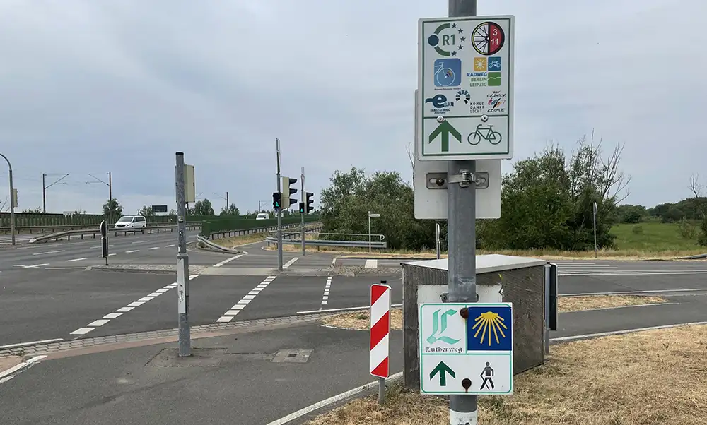 Am Rande einer großen Kreuzung steht ein Metallpfahl mit vielen, vielen Hinweisen auf Rad- und Wanderwege (Jakobsweg, Lutherweg)