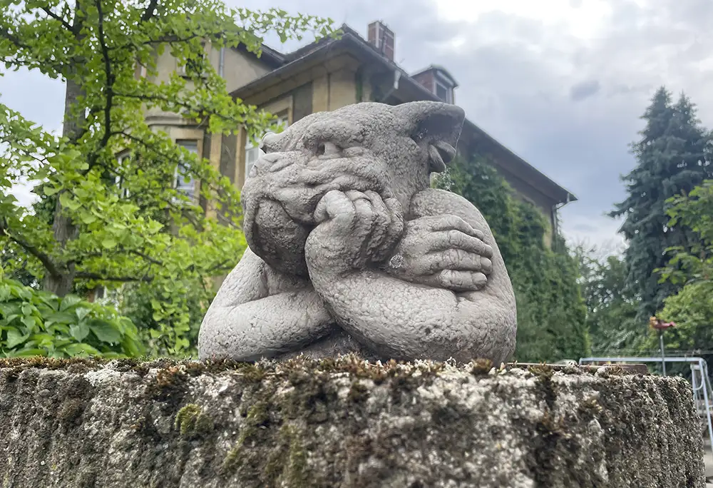 Stein-Skulptur eines schmollenden Gnoms mit Hundekopf auf einer Gartenmauer