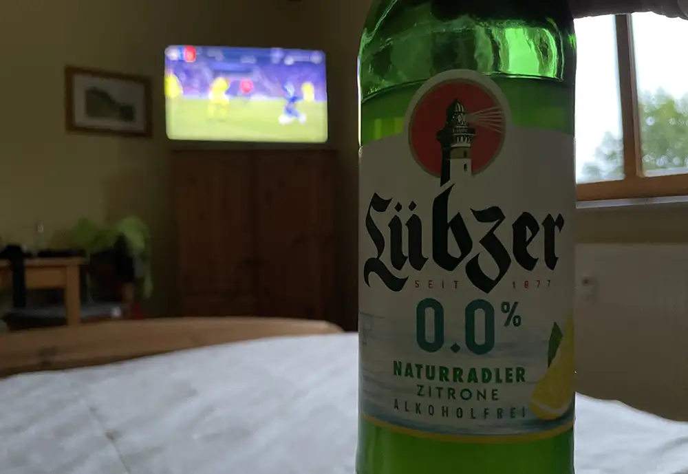 Flasche „Lübzer 0% Naturradler“ im dunklen Vordergrund, im Hintergrund unscharf ein Fußballspiel im TV