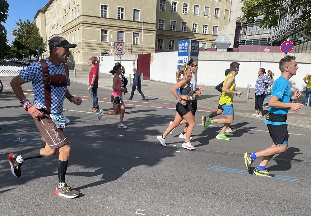 Große Säule mit „km 32“ am Straßenrand, davor die Marathon-läufer*innen, einer davon im Bayern-Outfit
