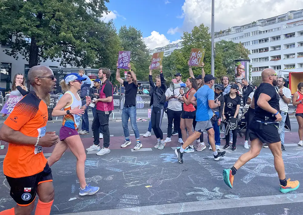 Jubelnde Adidas Runners halten am Streckenrand Plakate hoch, u.a. mit „You go girl!“