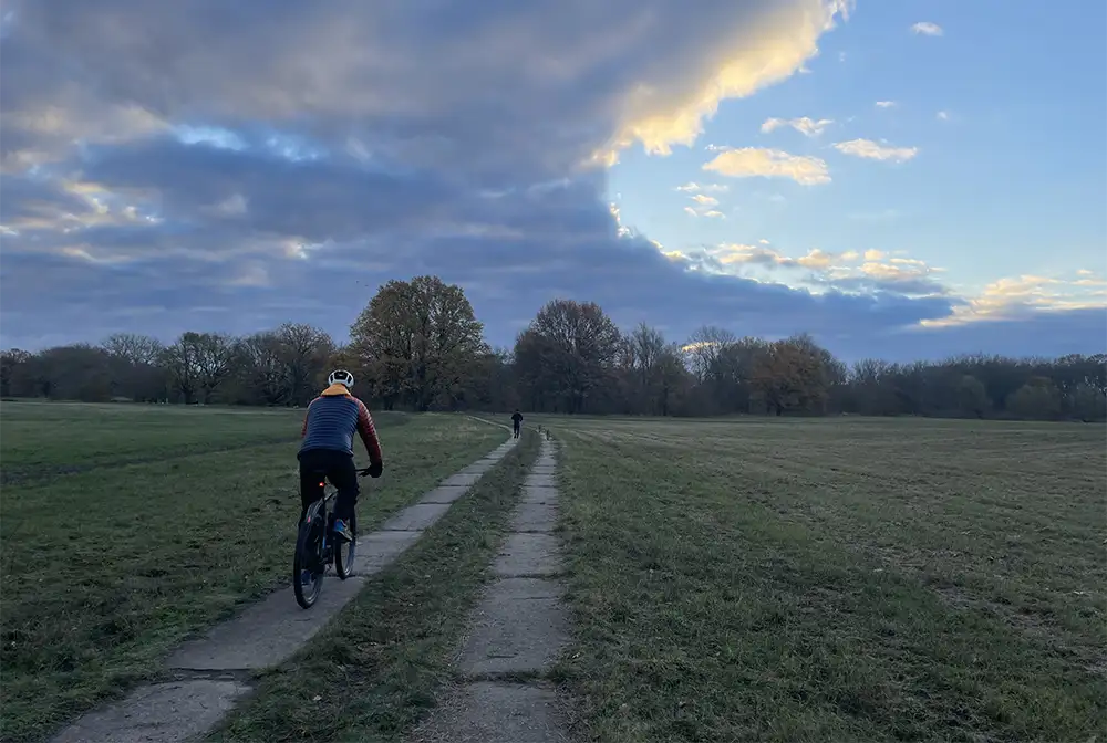 Radfahrer im Vordergrund und Läufer weit entfernt im Hintergrund auf Plattenweg zwischen Wiesen unter einem wolkigen Himmel