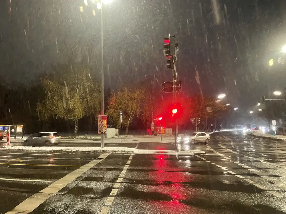 Große Straßenkreuzung im Morgendunkel mit Pfützen und sichtbarem Regen, die Fußgängerampel zeigt Rot