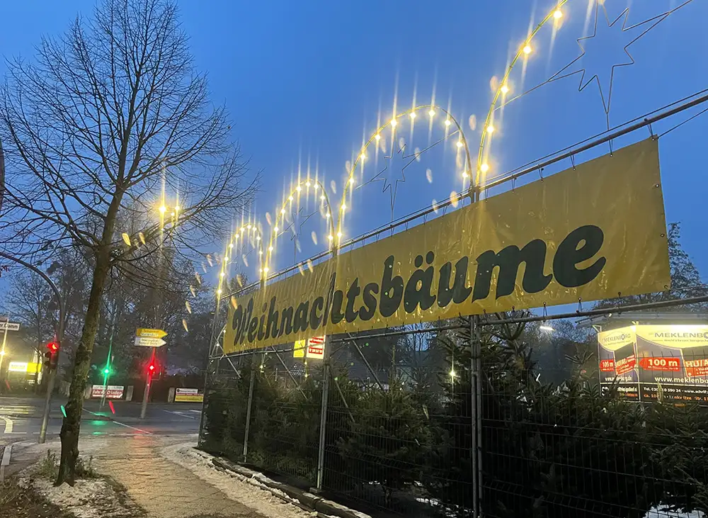 Großes gelbes Banner „Weihnachtsbäume“, darüber Bögen mit leuchtenden Glühlampen, an einem Weihnachtsbaumverkauf