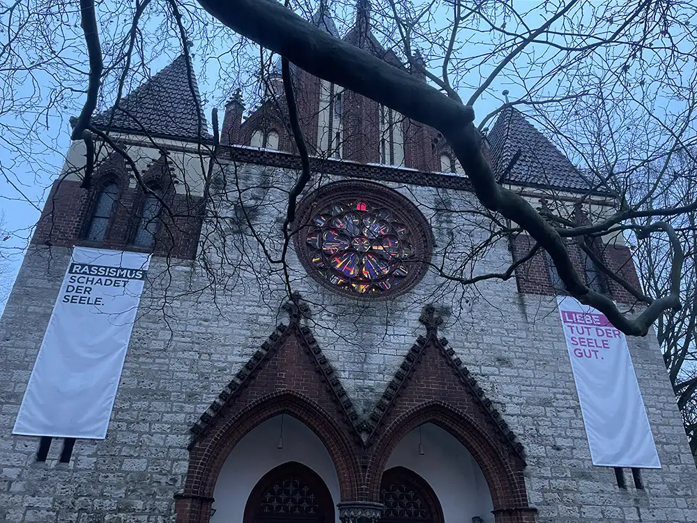 Große Kirche mit bunter Glasrosette an der Fassade, rechts und links hängen Banner: „Rassismus schadet der Seele“ und „Liebe tut der Seele gut“