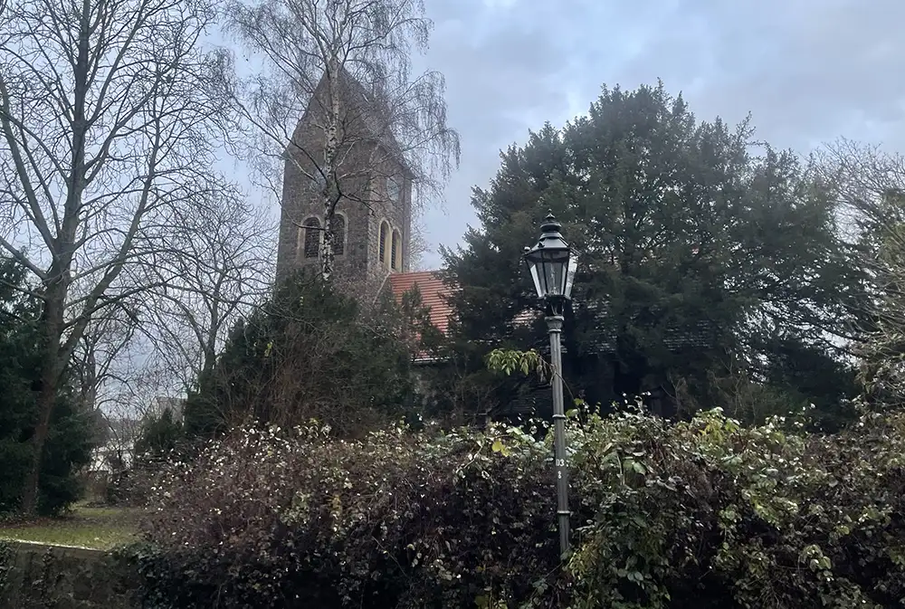 Alte Dorfkirche Lichtenrade, davor Büsche und Bäume und eine historische Laterne
