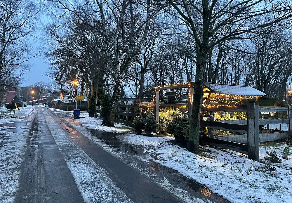 Schneematschige Dorfstraße im Morgengrauen, rechts ein mit Weihnachtsbeleuchtung erhellter Stand