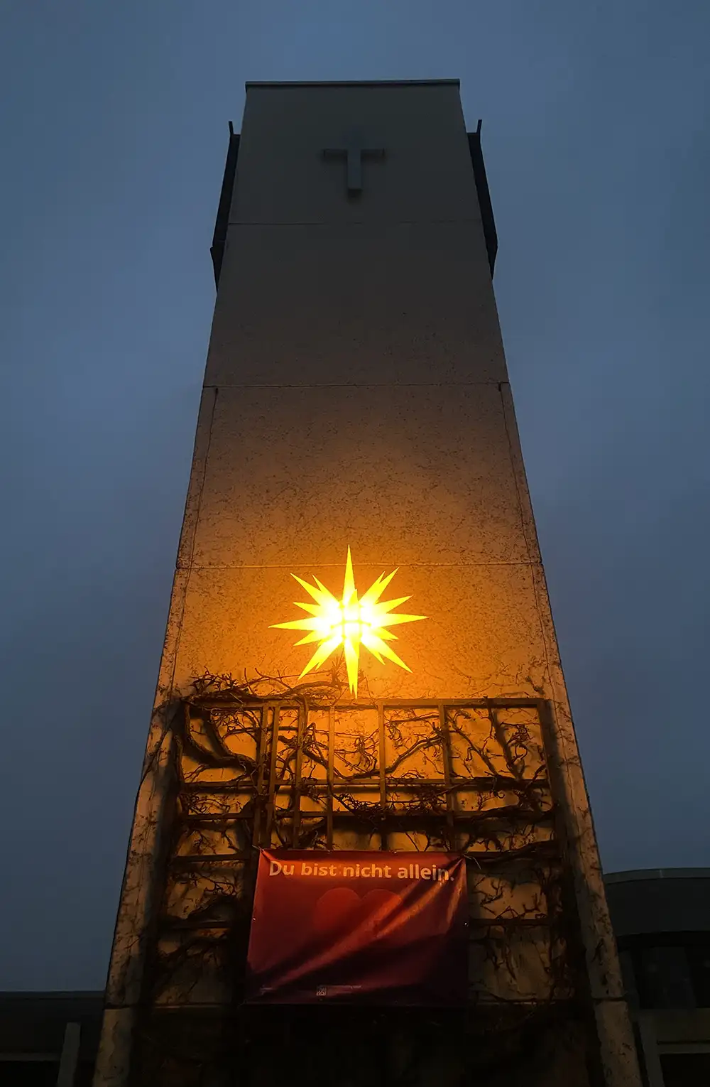 Kahler, moderner Kirch-Glockenturm mit großem gelb leuchtendem Hutschenreuther Stern und einem Banner „Du bist nicht allein.“