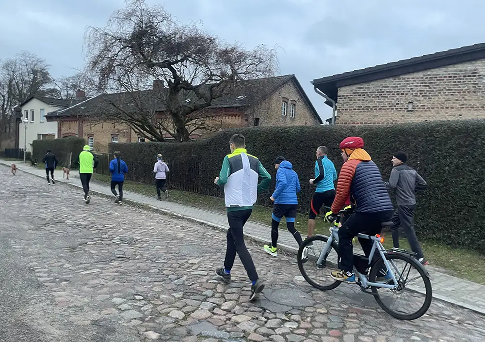 Laufgruppe mit acht Läuferinnen und Läufern sowie einem Radfahrer auf einer Kopfsteinpflasterstraße