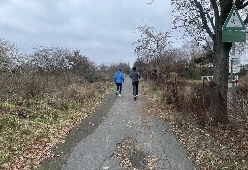 Zwei Läufer auf brüchigem Asphaltweg neben einer Kleingartenanlage