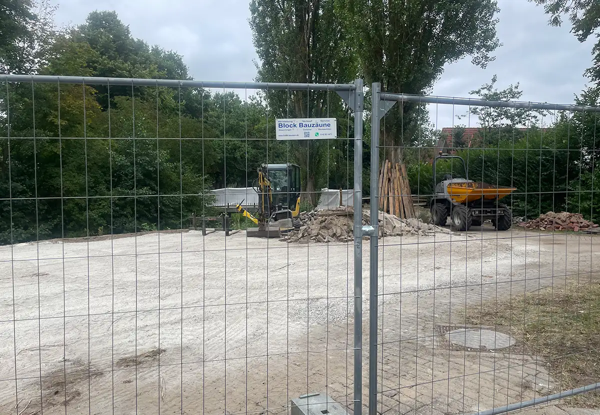 Hinter einem Gitter-Bauzaun sind zwei kleine Baufahrzeuge zu sehen