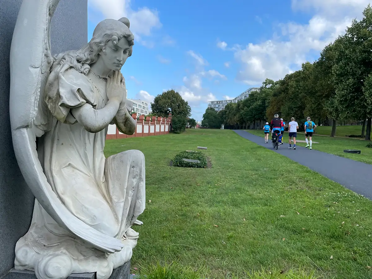 Weiße Marmorstatue eines Engels, im Hintergrund die Laufgruppe auf dem Asphaltweg durch den Friedhof