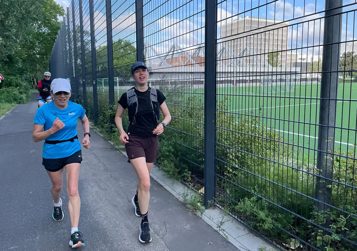 Zwei Läuferinnen laufen an einem hohen Metallzaun entlang, hinter dem ein Sportplatz liegt