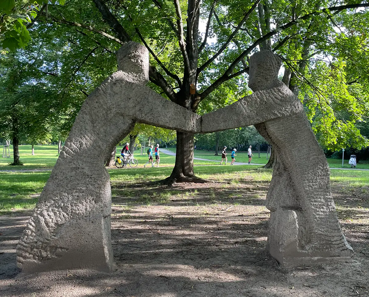 Denkmal „Wiedervereinigung“ in einem Park, zwei überlebensgroße Steinfiguren reichen sich die Hände und bilden einen Bogen, in dem man im Hintergrund die Laufgruppe sieht