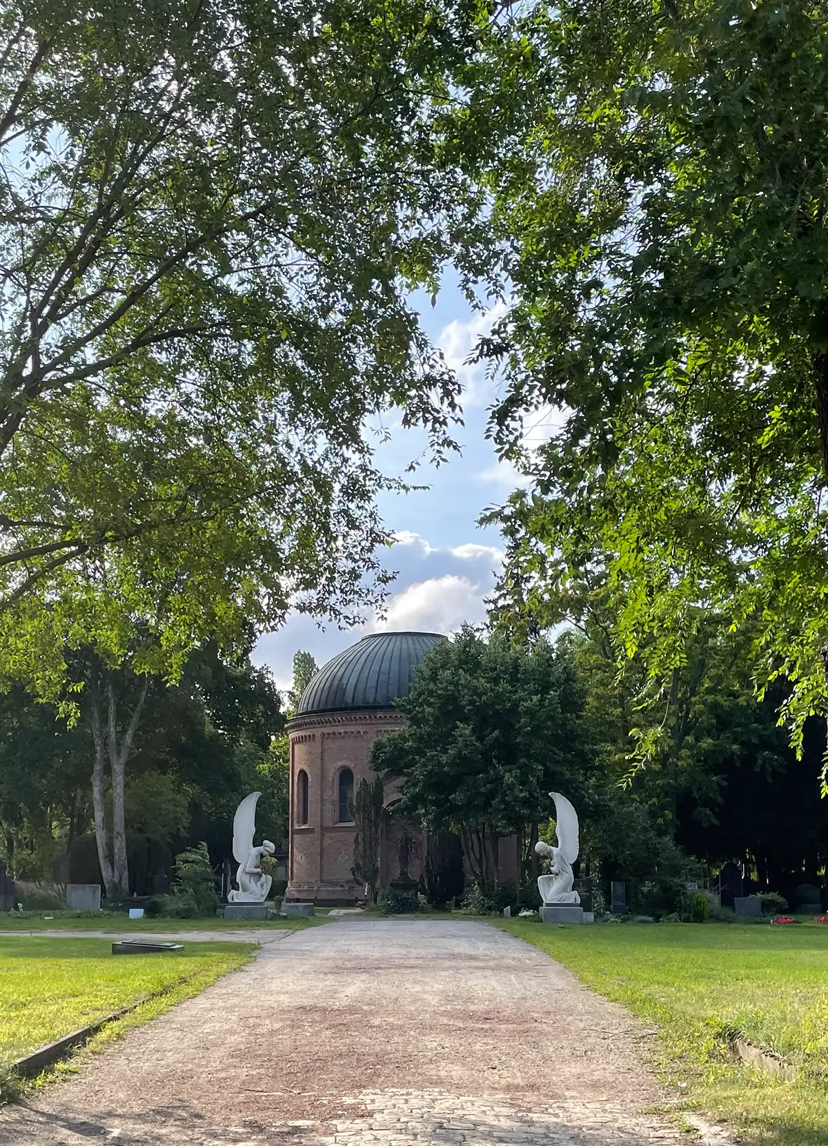 Breiter Weg zu einem Friedhofs-Gebäude,links und rechts säumen zwei überlebensgroße marmorne Engel den Weg