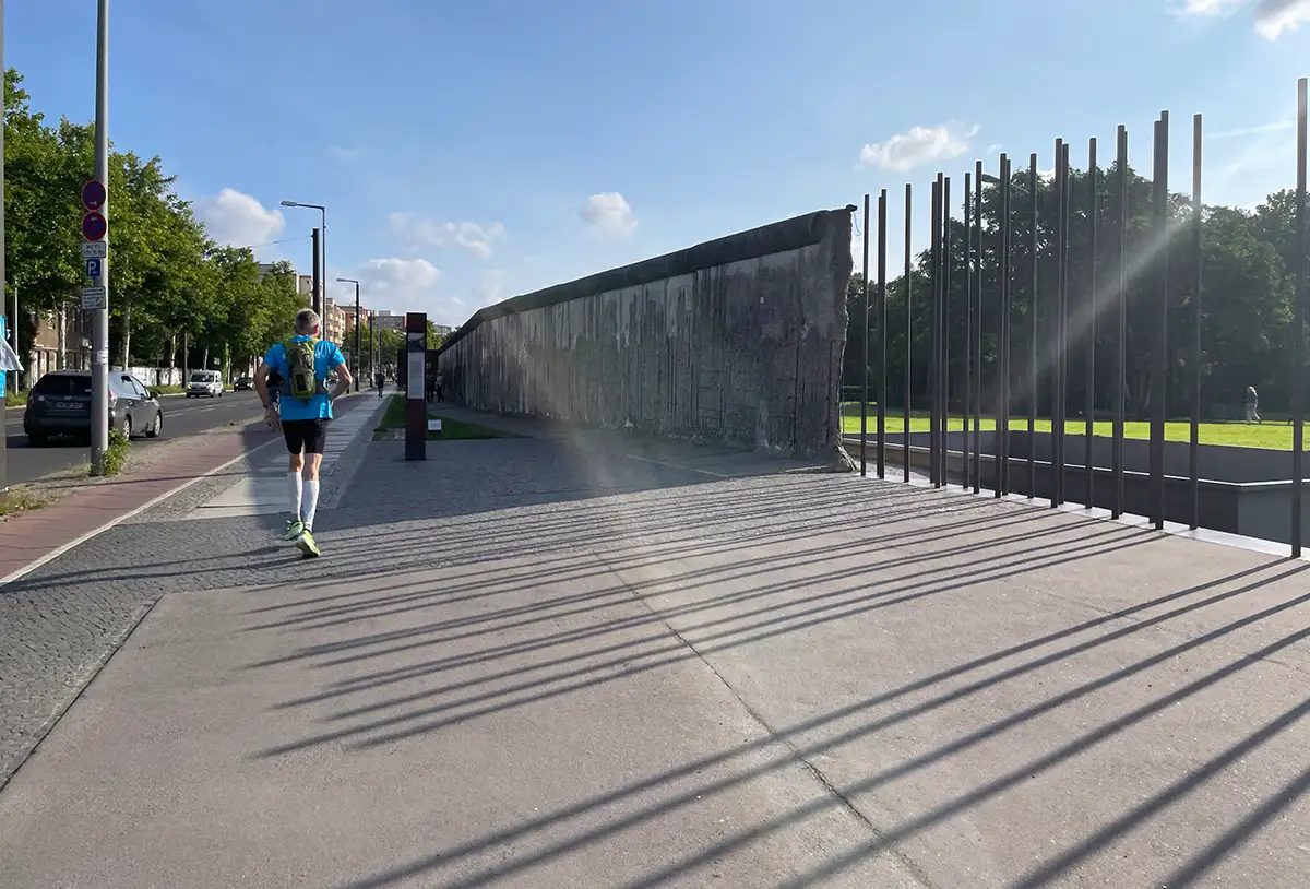Läufer neben Resten der Berliner Mauer, die von Metallstangen fortgeführt wird, die lange Schatten werfen