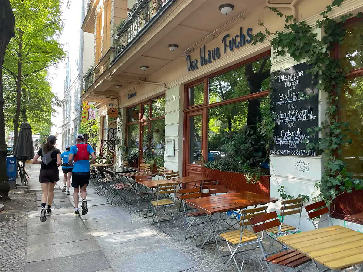 Laufgruppe kommt an einem Restaurant Der blaue Fuchs vorbei, vor dem Holztische und -stühle stehen